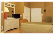 A simple bedroom with a bed, a dresser topped by a mirror and small TV, a lamp, and closed closet doors.
