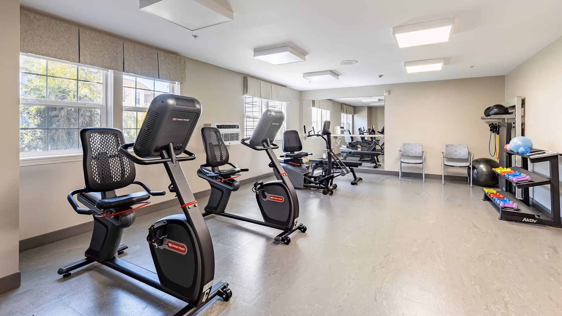 A bright fitness room with two stationary exercise bikes, a rowing machine, and various exercise equipment including stability balls, dumbbells, and resistance bands. The room has large windows with beige valances, a wall mirror, and two chairs against the far wall.