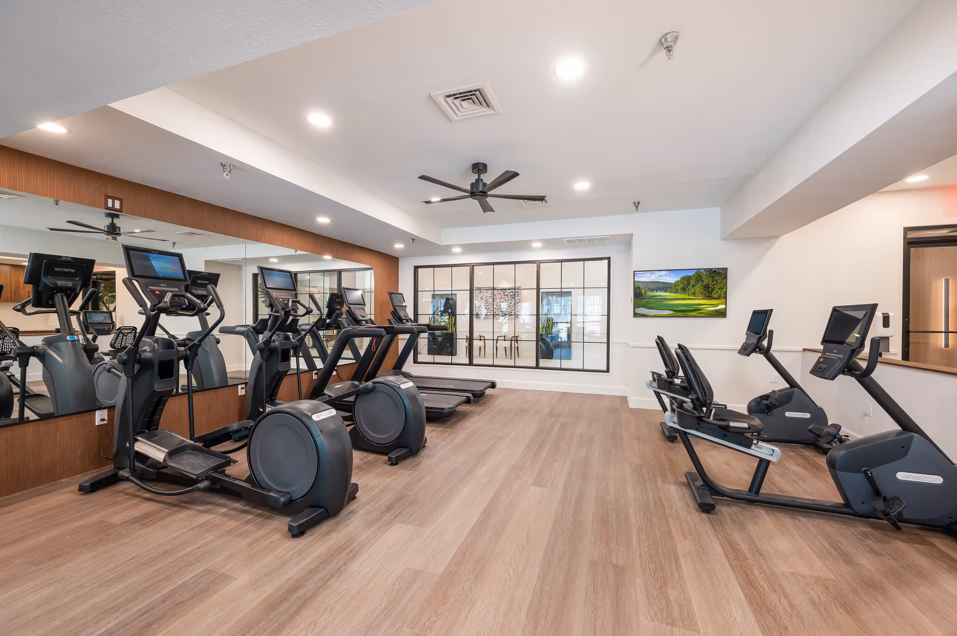 A modern fitness room with several cardio machines including treadmills, elliptical trainers, and recumbent bikes. The room has wood-style flooring, a large mirror on one wall, a ceiling fan, and a window looking into an adjacent room with chairs and artwork. A flat-screen TV is mounted on the wall.