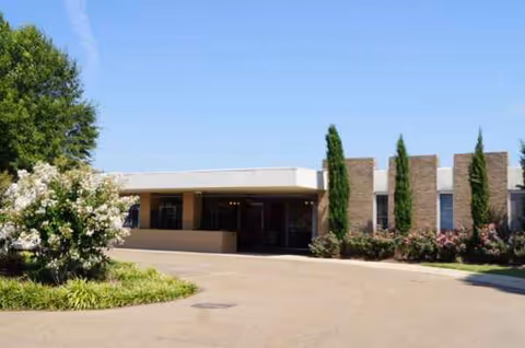 Front view of a single-story senior living facility with driveway, landscaping, and tall trees.