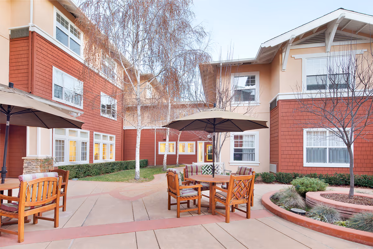 Outdoor courtyard area of a senior living facility with wooden chairs and tables under large umbrellas, surrounded by a two-story building with red and beige exterior walls, leafless trees, and some greenery.