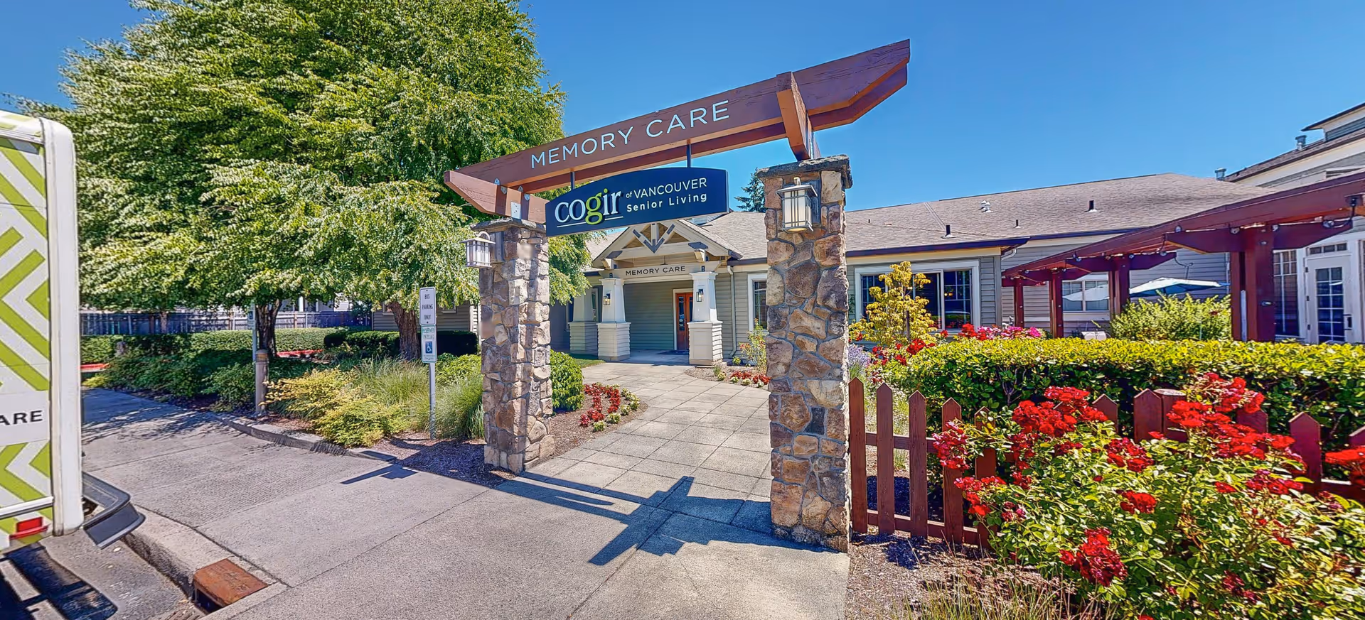 Entrance to a memory care facility with a stone archway labeled 'MEMORY CARE' and a sign for Cogir of Vancouver Senior Living. The pathway leads to a building surrounded by well-maintained landscaping with green bushes, red flowers, and trees under a clear blue sky.