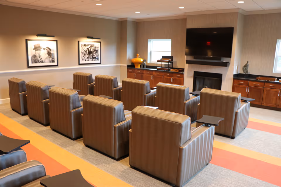 A cozy common area with multiple brown leather armchairs arranged in rows facing a wall-mounted flat screen TV above a fireplace. The room has warm lighting, framed black and white photographs on the wall, wooden cabinets with decorative items, and windows letting in natural light.