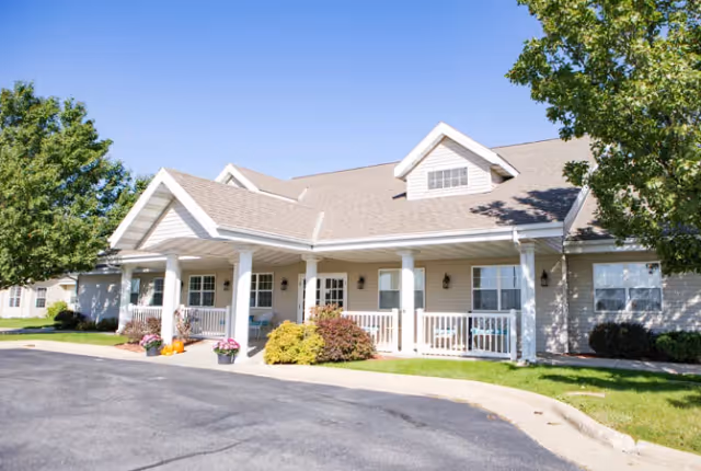 Exterior view of a single-story senior living facility building with a covered porch, white railings, and several windows. There are potted plants and shrubs near the entrance, and trees on either side of the building. The sky is clear and blue.