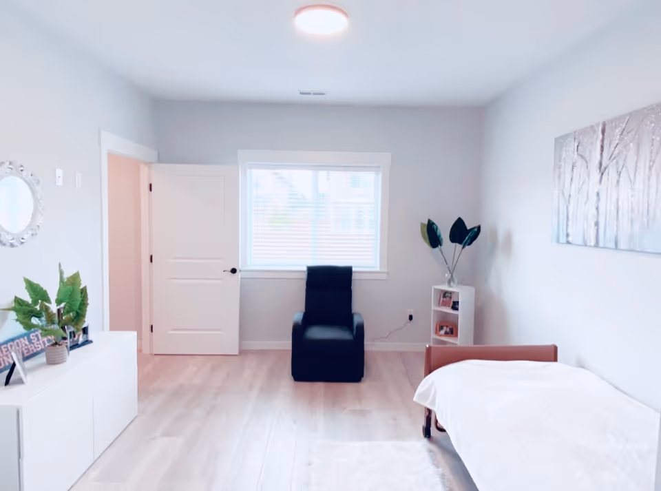 A bright and clean bedroom with light-colored walls and wooden flooring. The room features a single bed with a white blanket on the right side, a black armchair positioned in front of a window with blinds, a white cabinet with green plants and decorative items on the left, and a small white shelving unit with plants and framed photos near the bed. A painting of trees hangs on the wall above the bed.