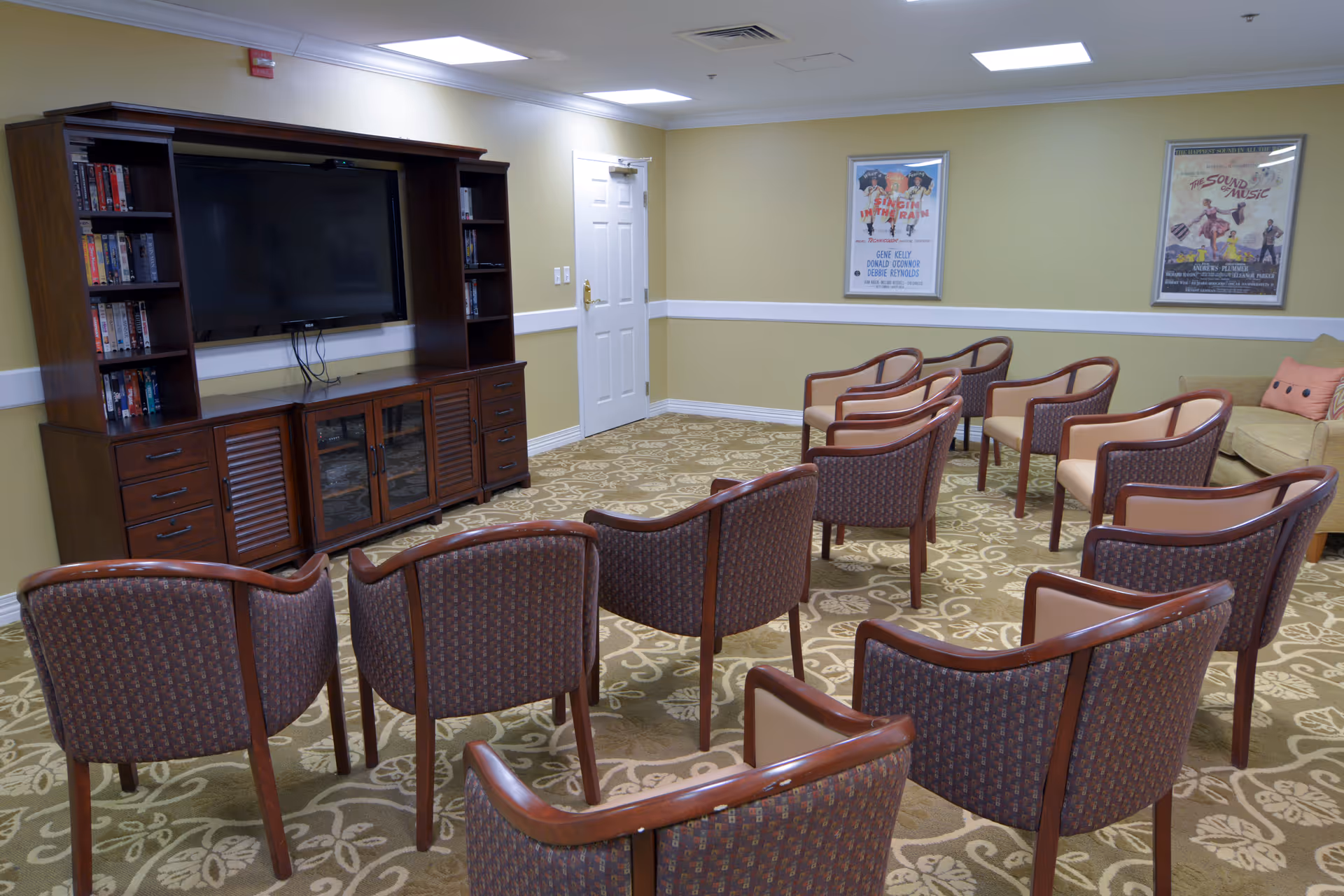Carpeted common room with rows of chairs facing a large TV and wooden entertainment cabinet, with framed posters on the walls.