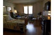 A senior care facility room with a bed, recliner, wheelchair, TV, and windows letting in natural light.