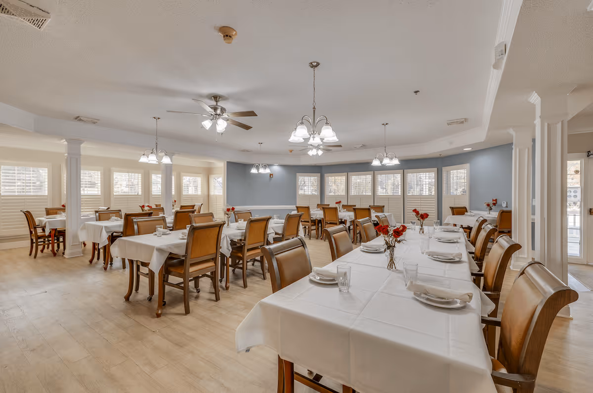 A spacious dining room with multiple tables covered in white tablecloths, each set with plates, glasses, and napkins. The room features wooden chairs with cushioned seats, large windows with white shutters, ceiling fans, and hanging light fixtures. Small vases with red flowers decorate the tables.