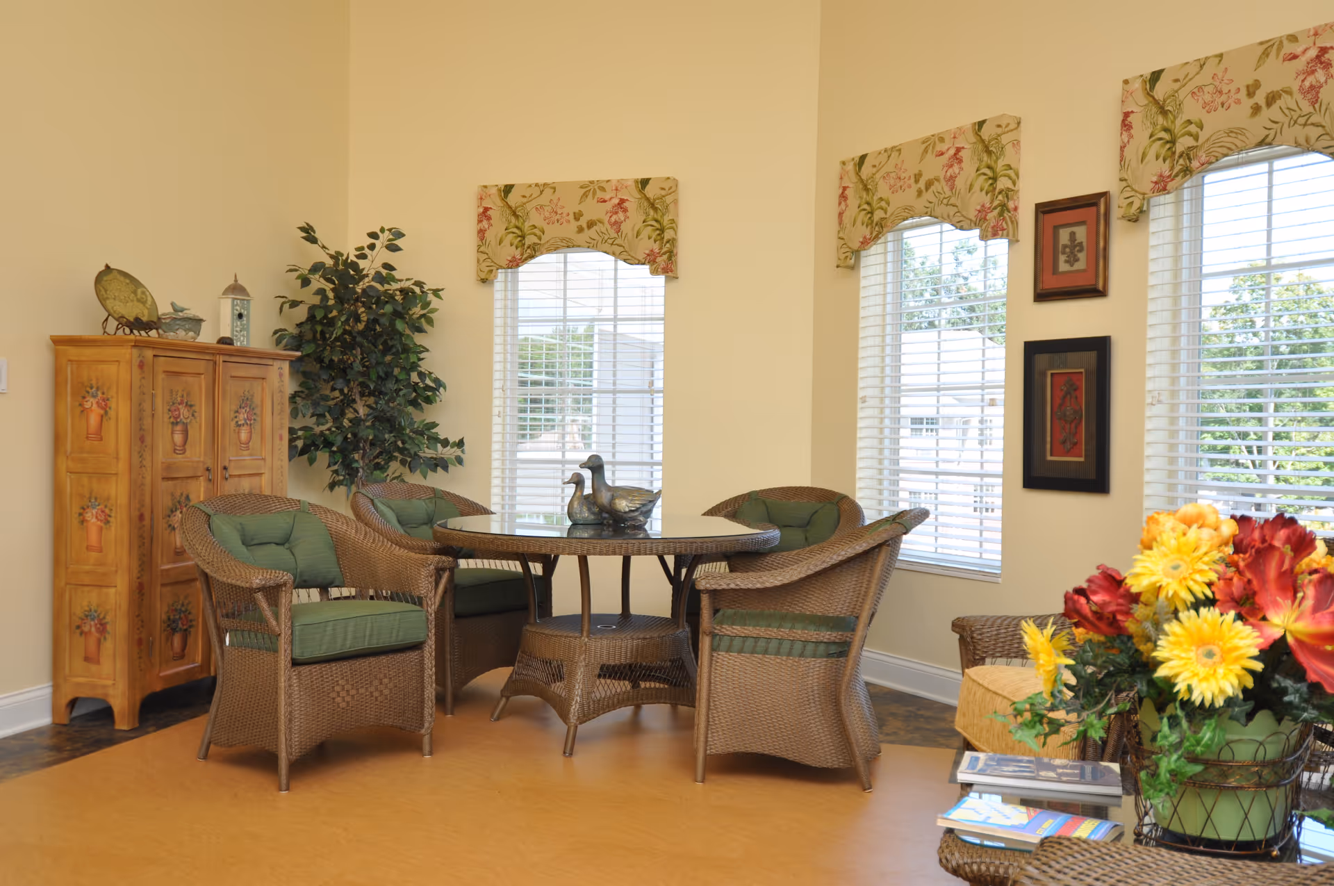 A cozy seating area with a round glass-top wicker table surrounded by four wicker chairs with green cushions. Behind the table is a wooden cabinet with floral designs and a tall artificial plant. The room has three large windows with floral valances, letting in natural light. On the right side, there is a wicker side table with a colorful flower arrangement and some magazines.