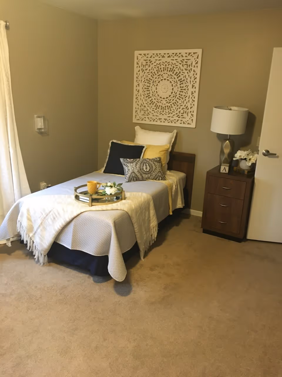 A neatly made single bed with white, yellow, and patterned pillows, a white blanket, and a tray with a yellow cup and flowers on top. Next to the bed is a wooden nightstand with three drawers, a modern lamp, a small decorative block with the letter 'A', and a vase with white flowers. The walls are painted beige, and there is a decorative white wall art piece above the bed. A white curtain is partially visible on the left side of the image.