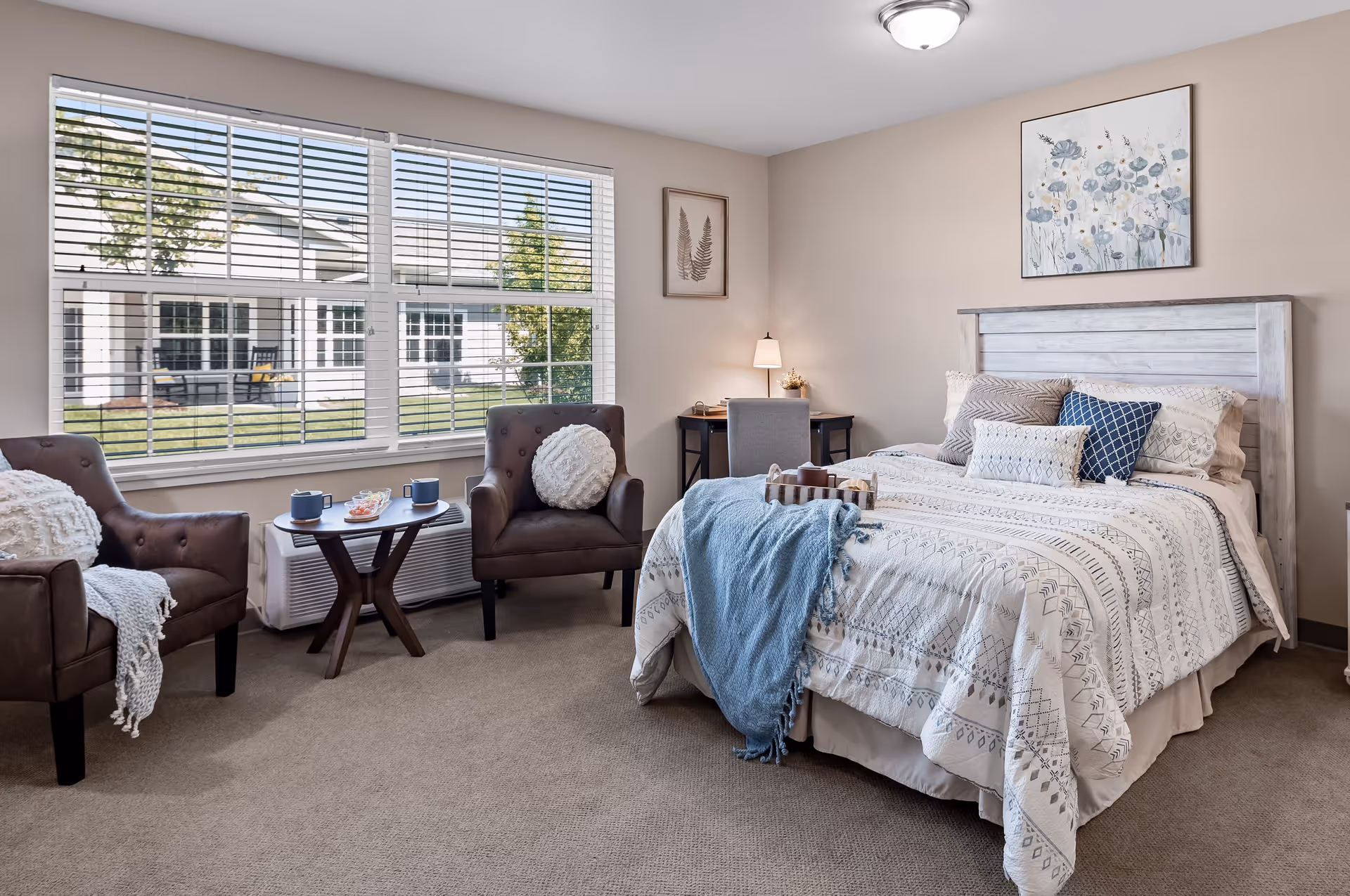 A cozy bedroom with a large window showing an outdoor view. The room features a bed with patterned bedding and multiple pillows, a blue throw blanket, two brown armchairs with white cushions, a small round wooden table with cups and snacks, a desk with a lamp and chair, and framed artwork on the walls.