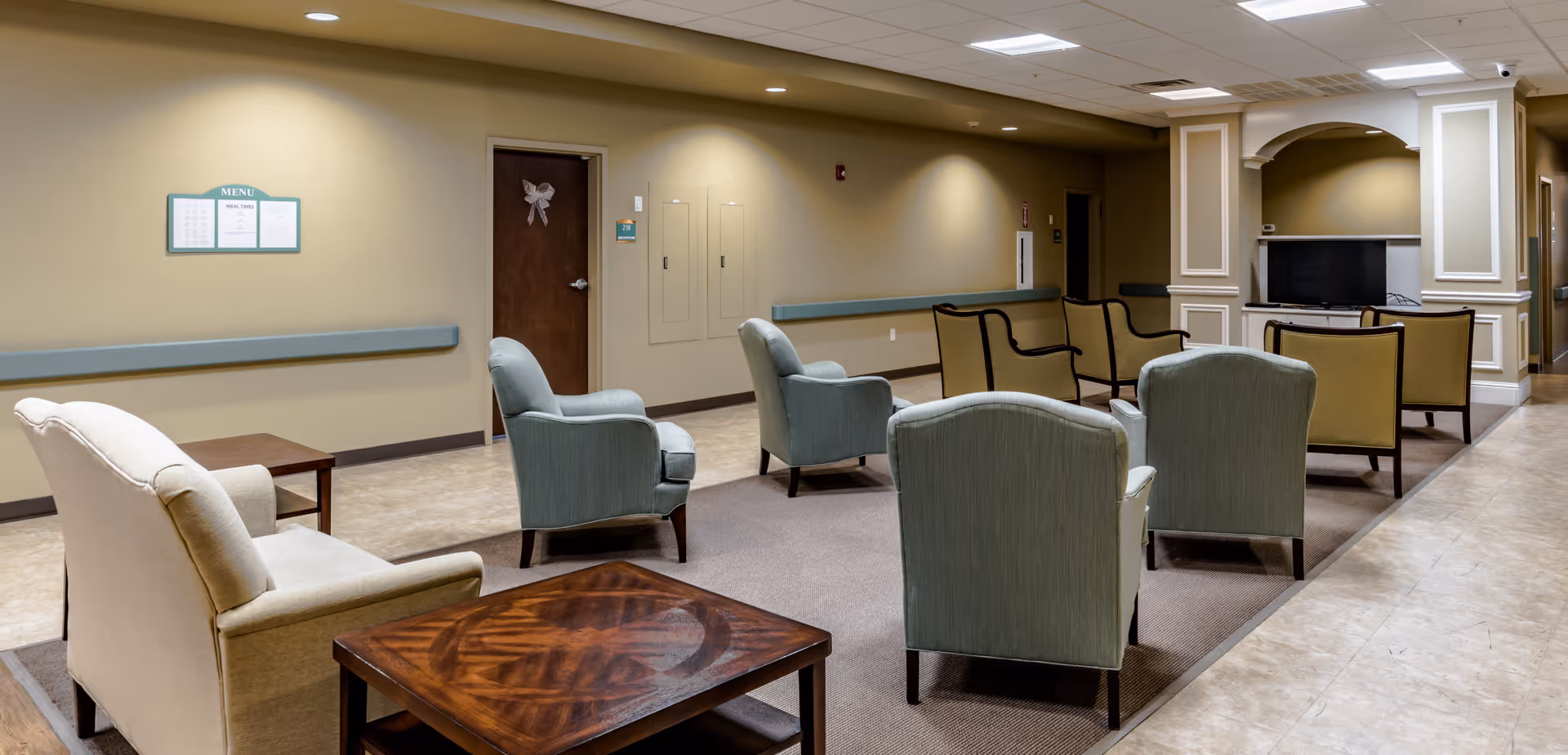 A seating area in a senior living facility with multiple upholstered armchairs arranged in rows facing a television mounted in a recessed wall niche. The room has beige walls, a tiled floor, and overhead fluorescent lighting. There is a wooden side table in the foreground and a door with a decorative bow on it in the background.