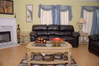 A cozy living room with a black leather sofa and matching armchair, a beige ornate coffee table with decorative bowls on top, light wood flooring, a fireplace with a white mantel on the left, and windows with blue and white curtains in the background.