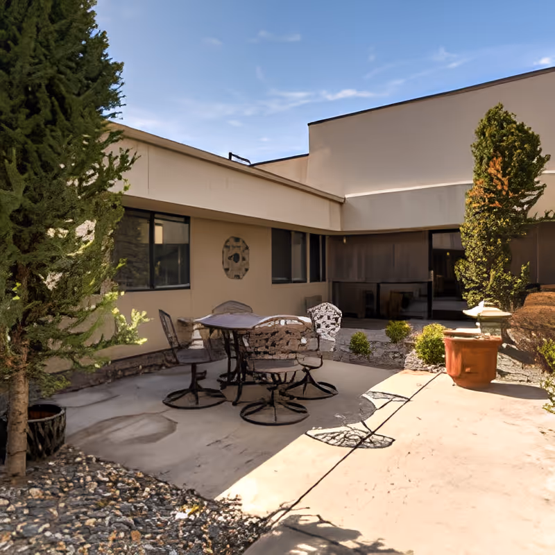 Outdoor courtyard patio with a metal table and chairs next to a low building, potted plants, and small trees.
