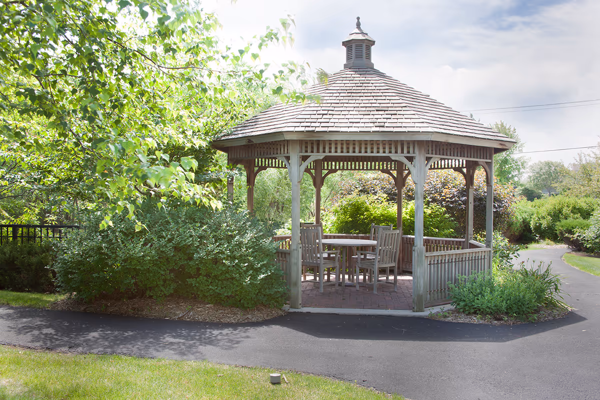 A wooden gazebo with a shingled roof situated in a garden area surrounded by green bushes and trees, with a paved walkway curving around it. Inside the gazebo, there is a round table with four wooden chairs.