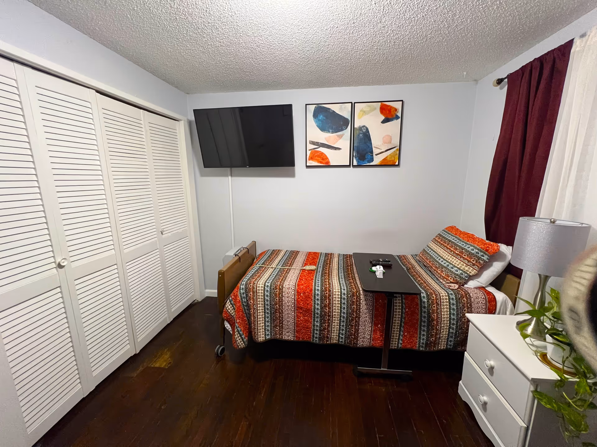 Small furnished bedroom with a single bed covered in a colorful striped quilt, wall-mounted TV, louvered closet doors, and a nightstand with a lamp.