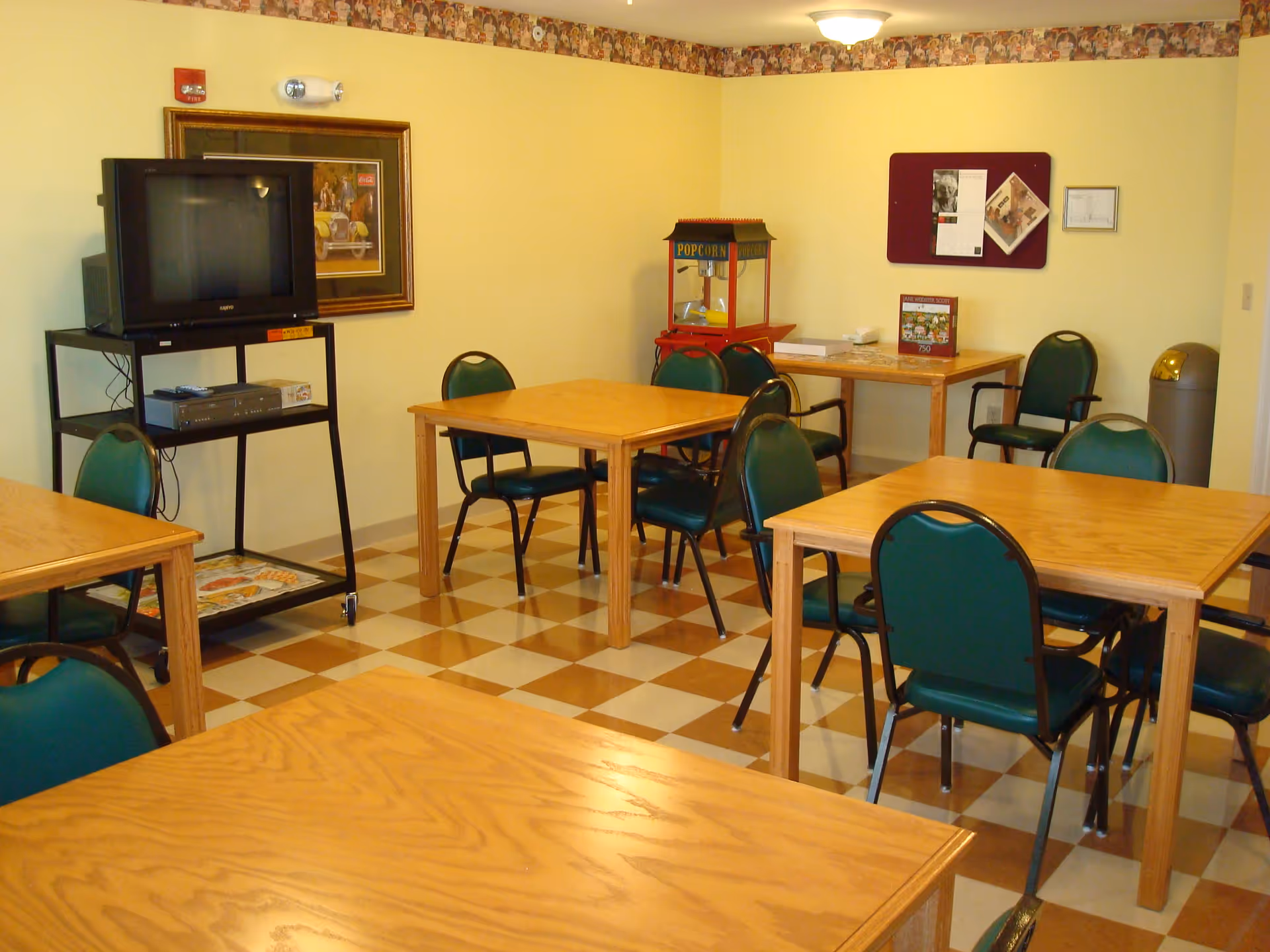 A small common room with several wooden tables and green cushioned chairs arranged around them. There is a popcorn machine in the corner, a TV on a black stand with a DVD player underneath, a framed picture on the wall, and a bulletin board with papers pinned to it. The floor has a checkered pattern with brown and beige tiles, and the walls are painted light yellow with a decorative border near the ceiling.