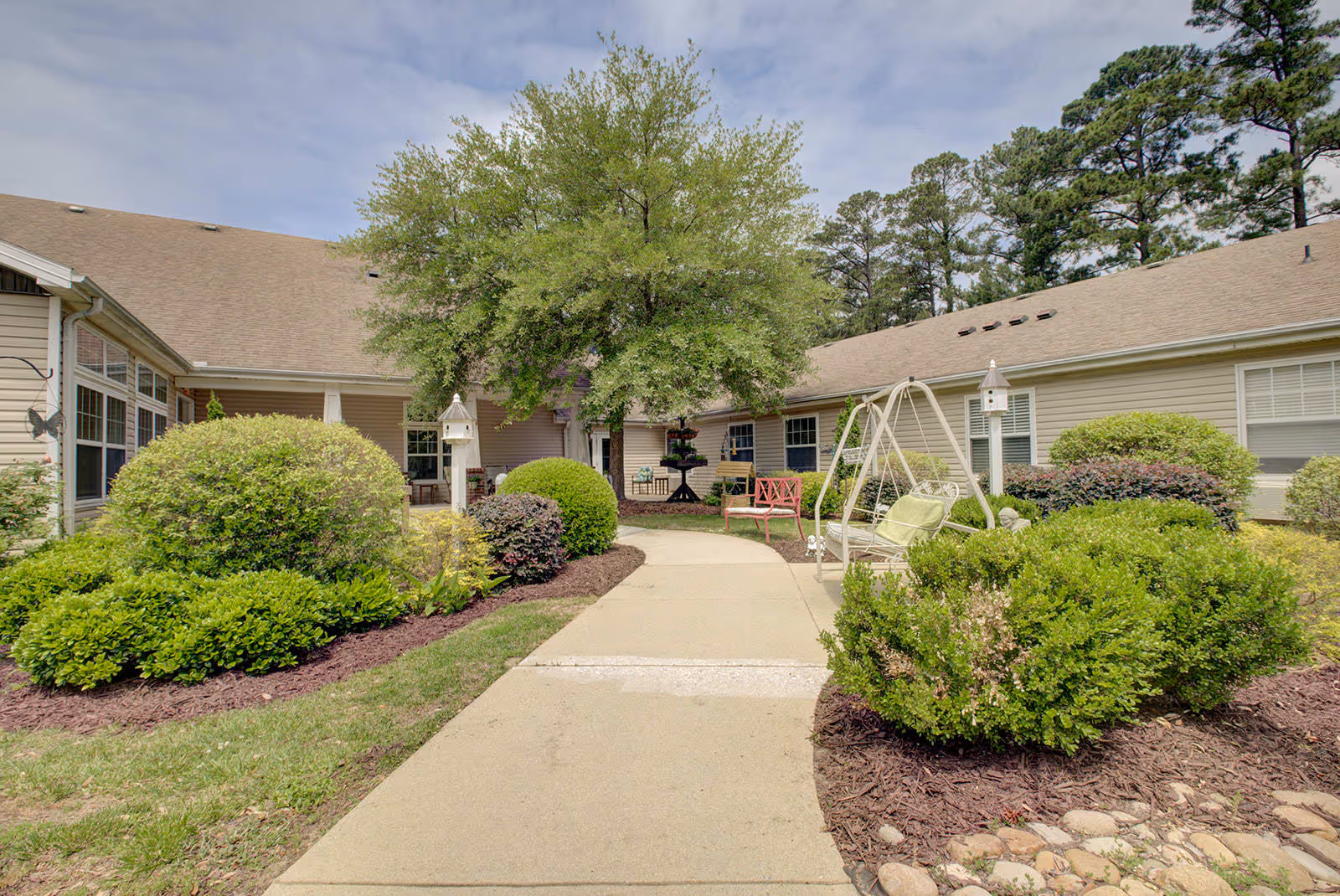 Outdoor courtyard area of Falls River Village Assisted Living featuring a paved walkway surrounded by well-maintained bushes and greenery, a large tree in the center, a white swing bench with green cushions, a red bench, and a multi-tiered black fountain near the building entrance.