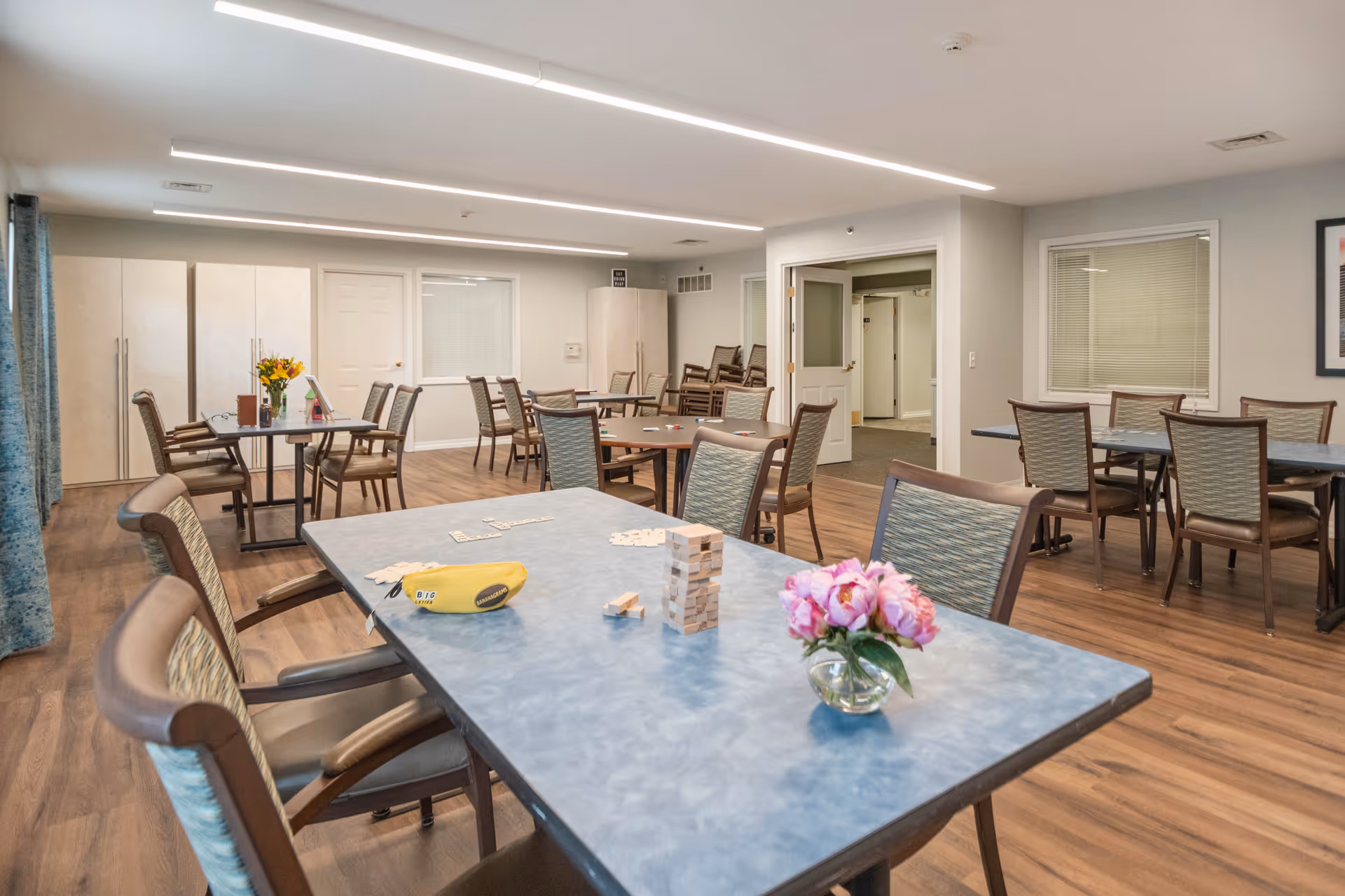 A spacious room with multiple tables and chairs arranged for group activities or dining. On the nearest table, there is a small vase with pink flowers, a Jenga game, and a yellow pouch. The room has wood flooring, light-colored walls, and overhead linear lighting. There are cabinets and a door along the back wall, and a window with blinds on the right wall.