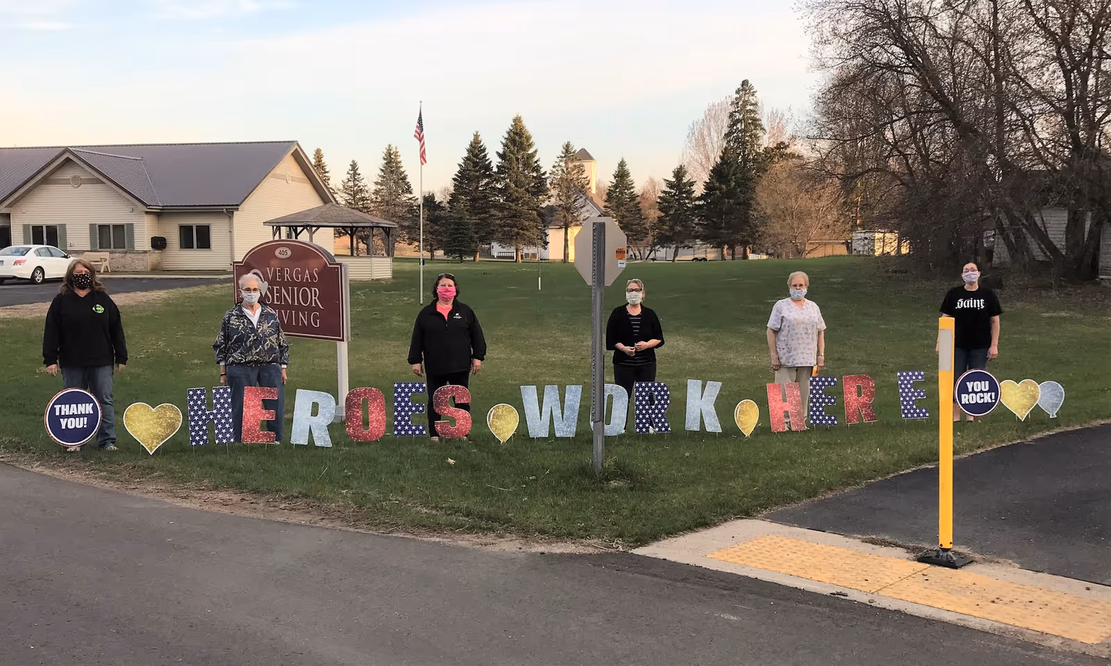 Six people wearing masks standing outside on grass near a sign that reads 'Vergas Senior Living'. They are holding or standing behind large colorful letters and symbols that spell out 'HEROES WORK HERE' with hearts and balloons, along with circular signs that say 'THANK YOU!' and 'YOU ROCK!'. Trees and buildings are visible in the background.