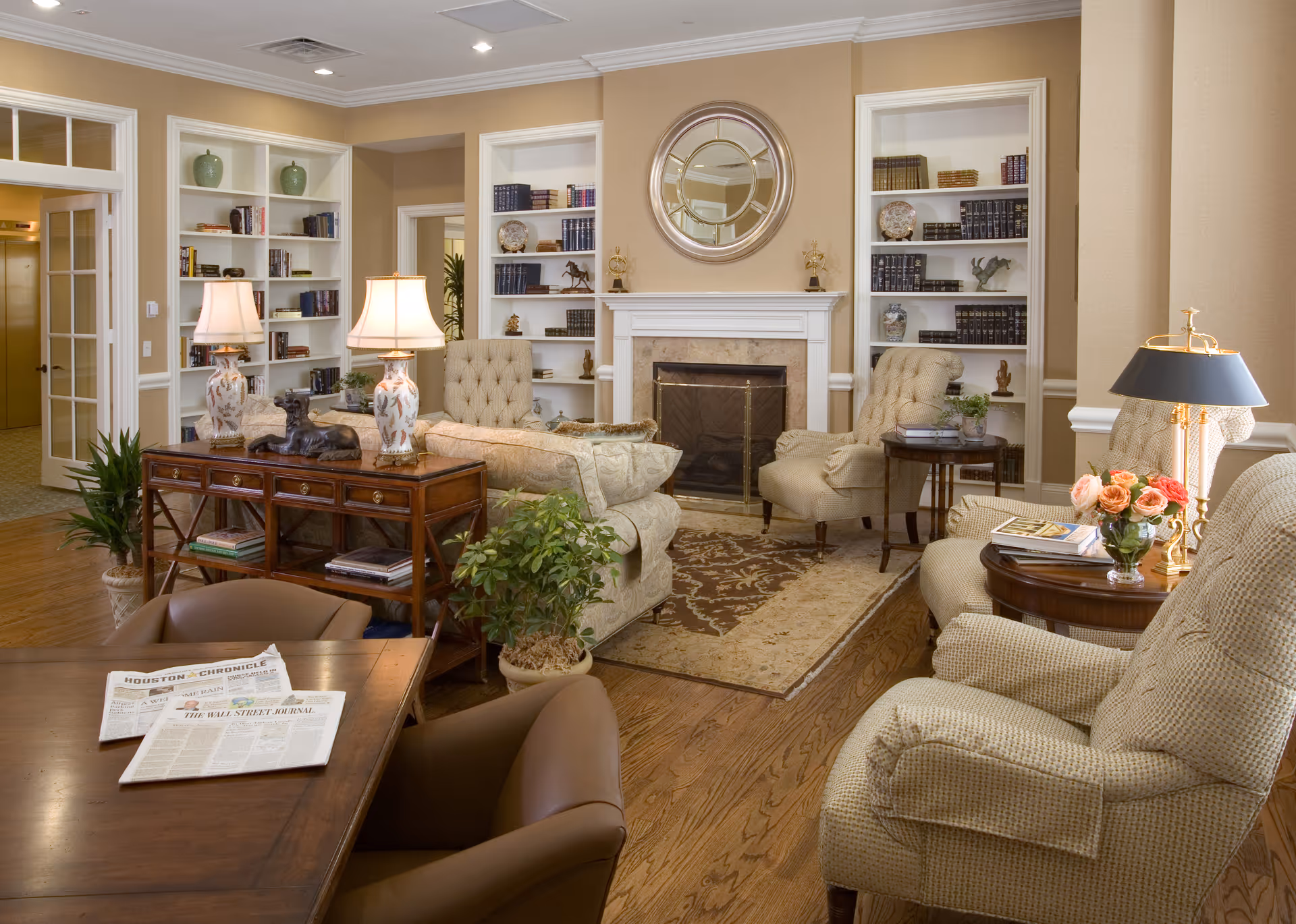 A cozy living room with beige walls and wooden flooring featuring a fireplace with a round mirror above it. The room has built-in white bookshelves filled with books and decorative items. There are several upholstered armchairs and sofas arranged around a patterned area rug. Two table lamps with floral bases sit on a wooden console table behind one sofa. A wooden table with newspapers and brown chairs is in the foreground, and a vase of pink roses is on a side table next to one armchair.