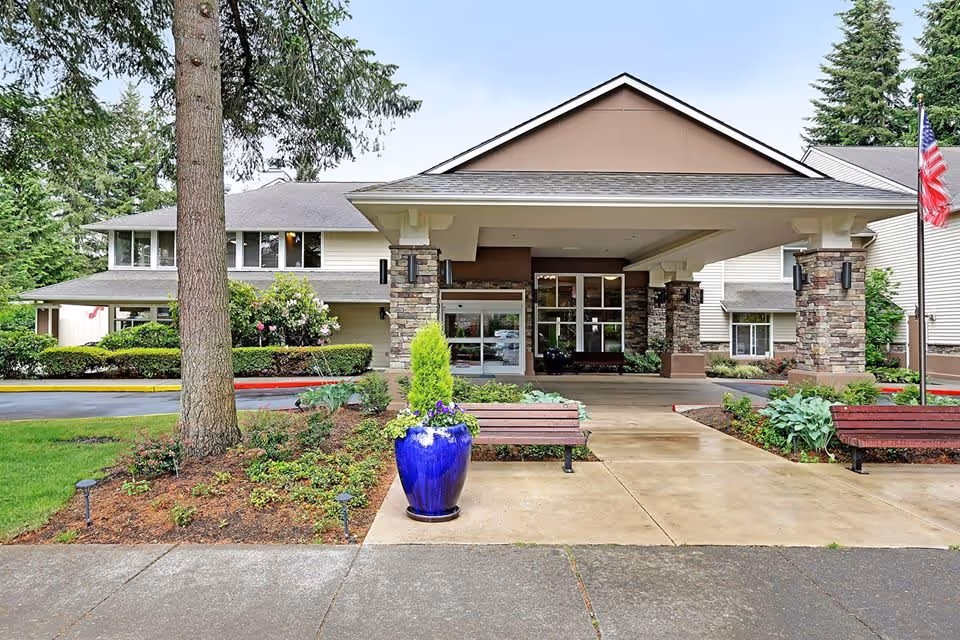 Front exterior view of Windsor Square Senior Living facility with a covered entrance supported by stone pillars, benches on either side of the walkway, a large blue planter with greenery, and an American flag on the right side. The building is surrounded by trees and landscaping.