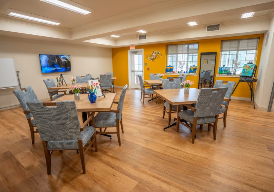 Bright common room with wooden tables and patterned chairs arranged for activities, a wall-mounted TV, and a yellow accent wall with windows.