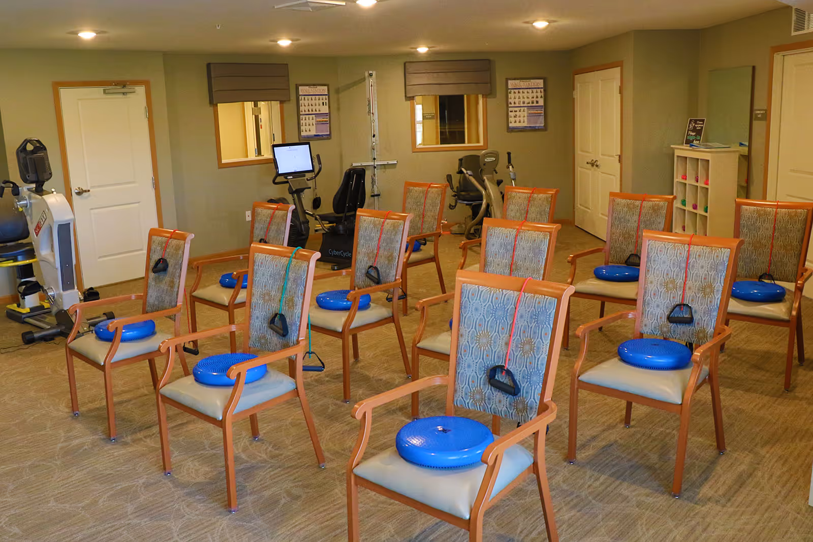 A senior living facility exercise room with multiple wooden chairs arranged in rows. Each chair has a blue balance cushion and resistance bands attached to the backrest. In the background, there are exercise machines and two windows with blinds. The room has beige walls and carpeted flooring.