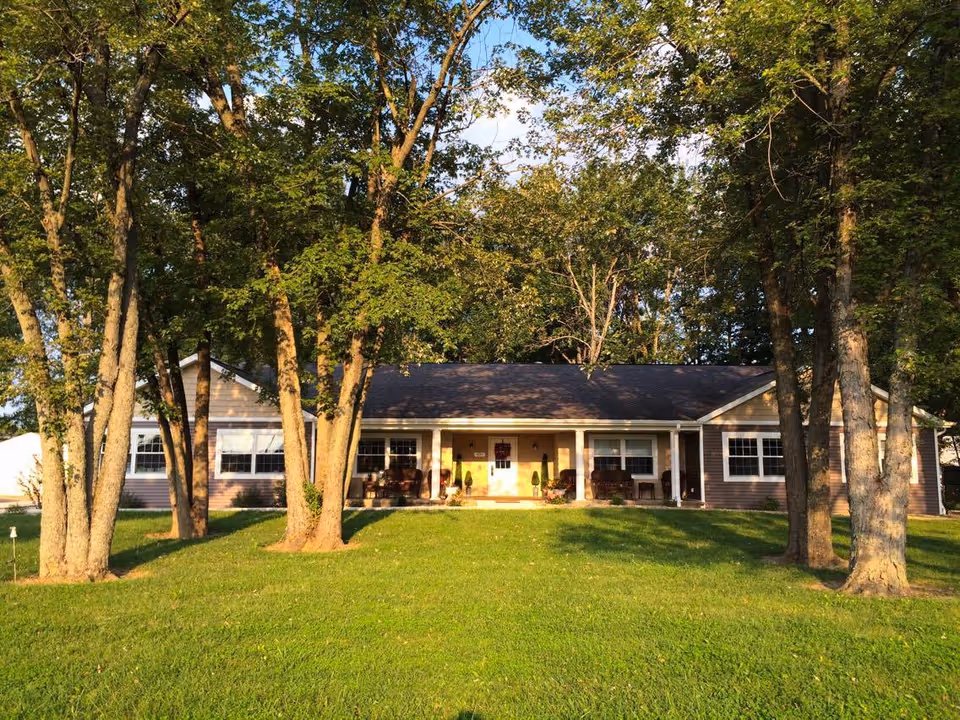 Single-story residential building with a covered front porch set behind a grassy lawn and shaded by several trees.