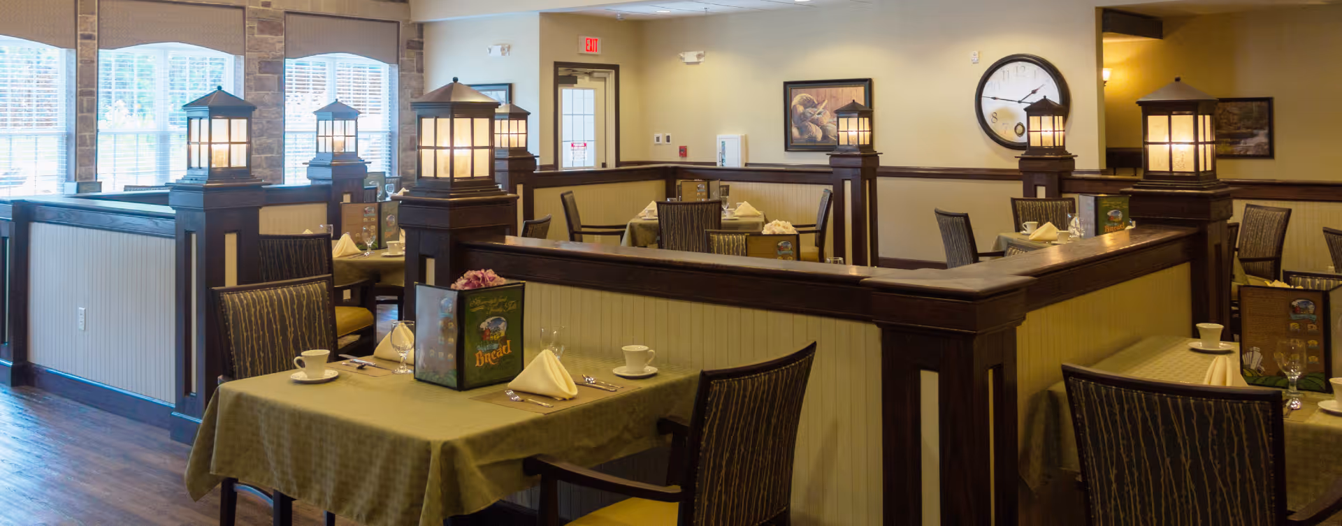 Interior view of a dining room in a senior living facility with tables covered in green tablecloths, set with cups, napkins, and utensils. The room features wooden partitions with lantern-style lights on top, large windows with blinds, and framed artwork on the walls.