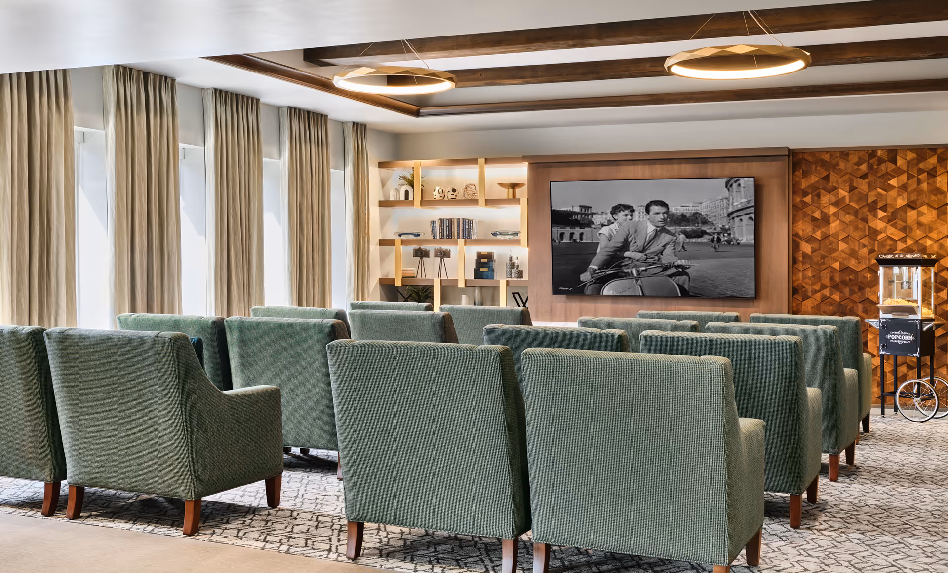 A cozy senior living facility media room with rows of green upholstered armchairs facing a large wall-mounted screen displaying a black and white movie scene. The room features large windows with beige curtains, wooden ceiling beams, modern circular ceiling lights, a popcorn machine on the right, and built-in shelves with decorative items and books.