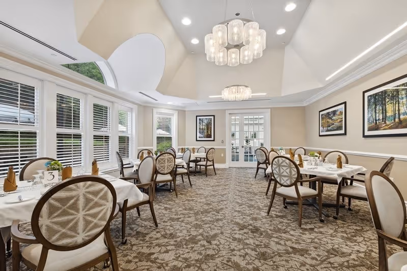 A bright and elegant dining room with multiple round tables covered with white tablecloths, each set with napkins, glasses, and small flower arrangements. The room features large windows with white blinds, beige walls adorned with framed landscape paintings, and two modern chandeliers hanging from a high, vaulted ceiling.
