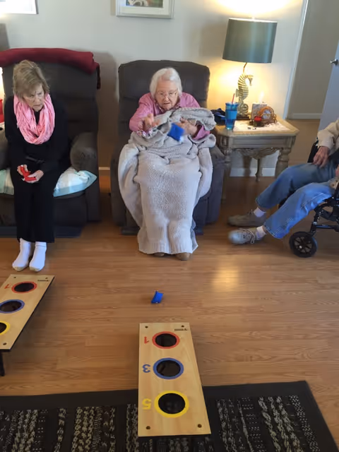 Three elderly individuals seated in a living room playing a bean bag toss game. One woman in the center is throwing a blue bean bag towards a wooden board with numbered holes. The room has wooden flooring, a patterned rug, and a side table with a lamp and some items on it.