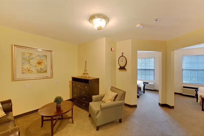 A cozy senior living room area with beige walls and carpet. The room features a brown leather sofa, a gray armchair with a patterned pillow, a wooden coffee table with a small green plant, and a dark wooden dresser with a decorative Eiffel Tower model. A framed floral painting hangs on the wall, and a round clock is mounted on a corner wall. Two adjacent rooms with large windows and beds are visible in the background.