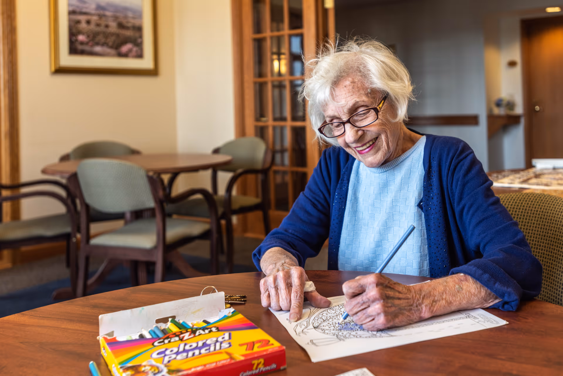 An elderly woman sitting at a table in a communal room, smiling as she colors with colored pencils.