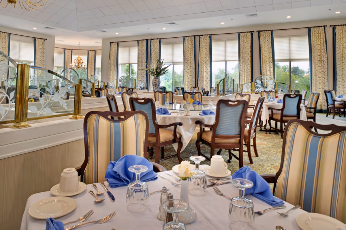 A bright and elegant dining room with round tables covered in white tablecloths, set with plates, glasses, silverware, and blue napkins. The chairs have wooden frames with striped upholstery. Large windows with patterned curtains allow natural light to fill the room. Decorative glass partitions with floral etching separate sections of the dining area.