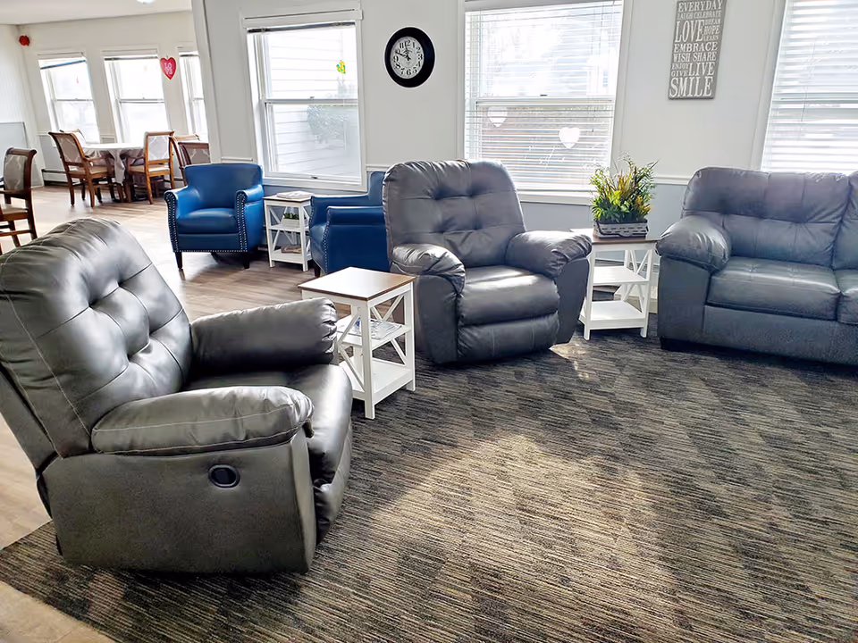 A bright and cozy living room area with three gray leather recliners arranged around two white side tables. Behind the recliners, there are two blue armchairs and a dining area with wooden chairs and tables near large windows letting in natural light. A clock and a decorative sign are mounted on the wall, and a potted plant sits on one of the side tables.