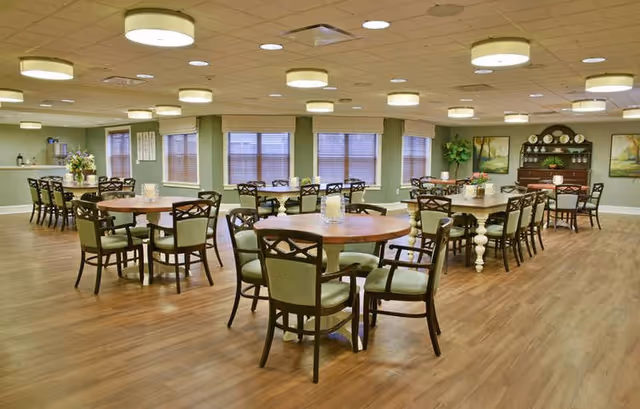 Spacious dining room with round wooden tables and green-upholstered chairs arranged on wood floors beneath round ceiling lights.