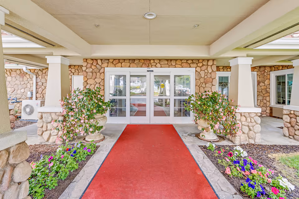 Entrance to a senior living facility with a red carpet leading to double glass doors. The exterior walls are made of stone, and there are two large planters with pink flowers on either side of the entrance. Flower beds with colorful flowers line the walkway.