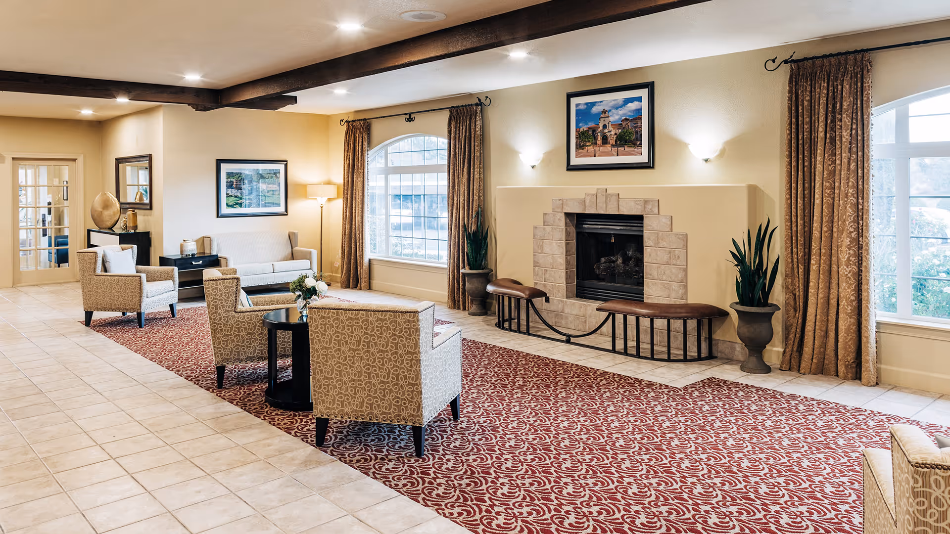 A bright communal living room with armchairs and sofas arranged on a patterned rug facing a fireplace beneath a framed painting.