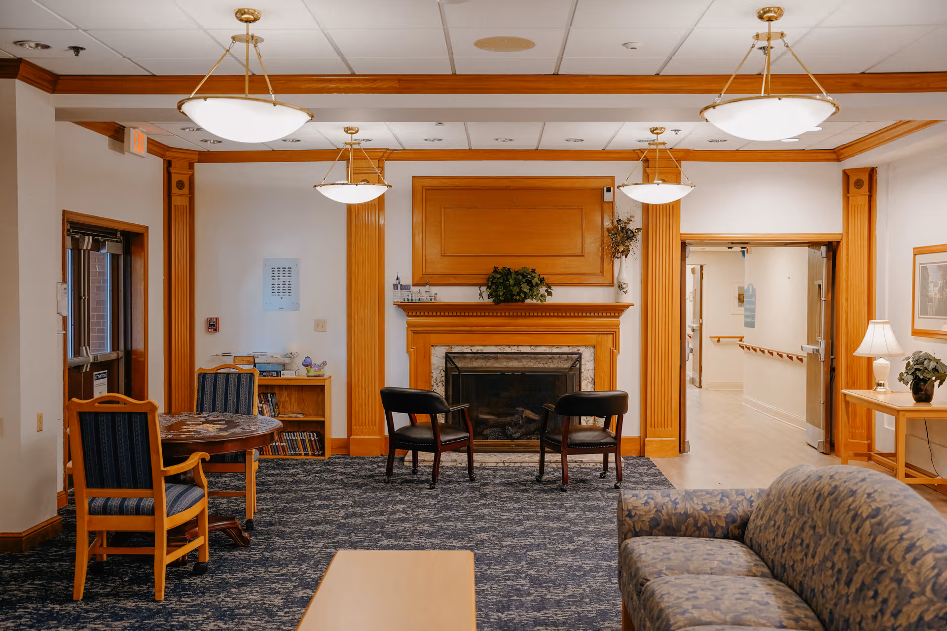A cozy communal lounge with a fireplace, chairs, a table, and a sofa in a senior living facility.
