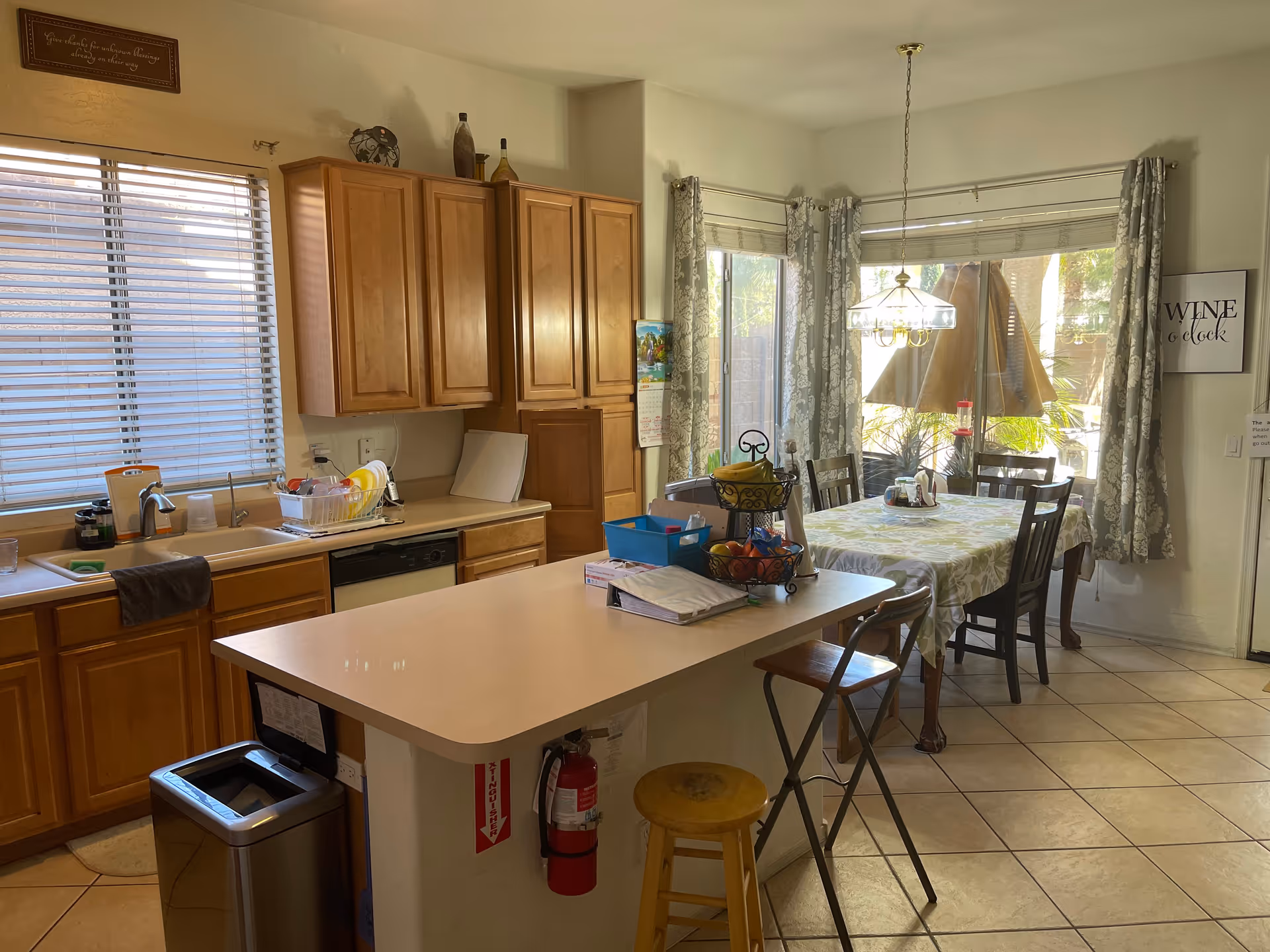 A bright kitchen and dining area with wooden cabinets, a sink with dishes drying on a rack, a kitchen island with stools, and a dining table covered with a floral tablecloth surrounded by chairs. Large windows with patterned curtains let in natural light, and there is a hanging light fixture above the dining table.