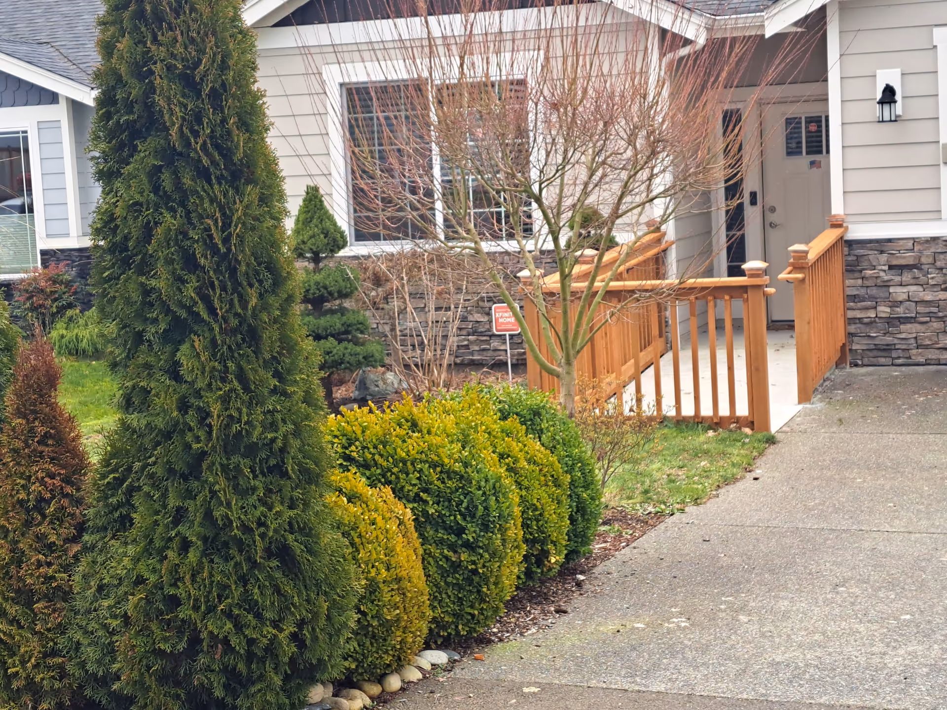 Front exterior view of a residential building with a concrete driveway, a wooden ramp with railings leading to the front door, and landscaped bushes and trees in the yard.