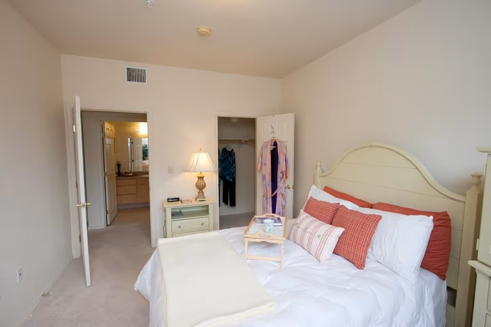 A bedroom with a cream-colored bed frame and white bedding accented with red and striped pillows. A small wooden tray with a cup and a book is placed on the bed. To the right of the bed is a nightstand with a lamp and some items on it. The room has beige carpet and walls. There is an open door leading to a bathroom and another open door revealing a closet with clothes hanging inside.
