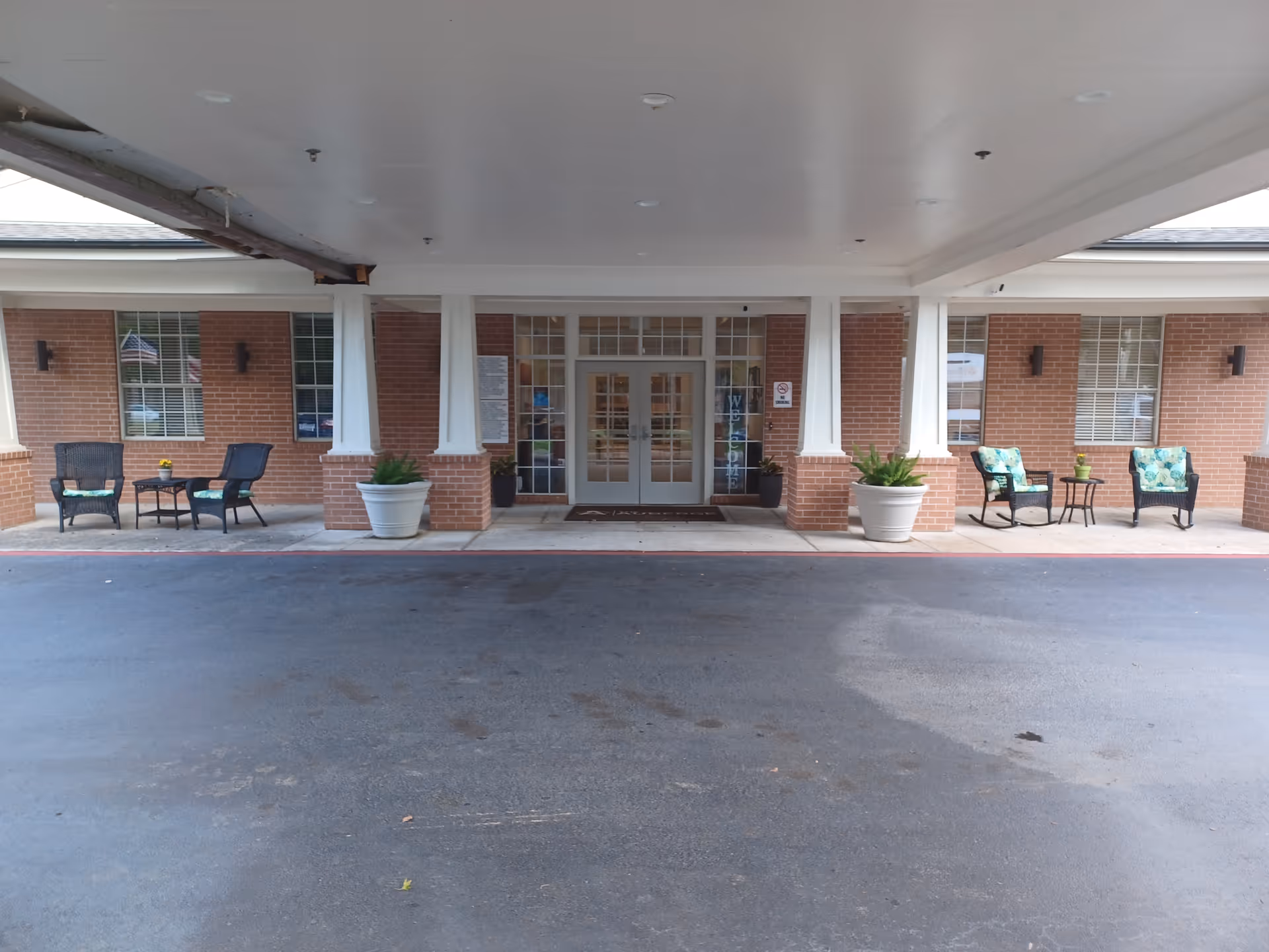 Covered entrance/portico of a brick senior living facility with double glass doors, columns, potted plants and outdoor seating.