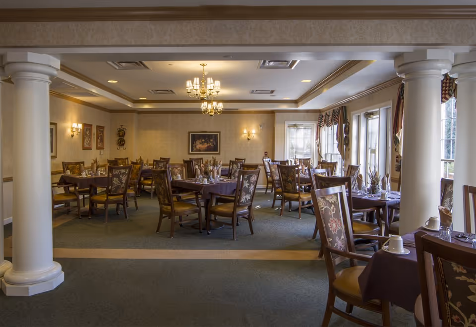 A dining room with multiple tables covered in dark tablecloths, each set with cups, glasses, and napkins. The room features upholstered wooden chairs with floral patterns, large white columns, chandeliers, wall sconces, framed artwork, and windows with decorative curtains allowing natural light to enter.