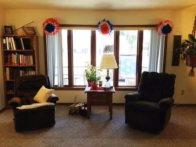 Two recliner chairs flank a small wooden side table with a lamp and plants in front of a large window decorated with red, white, and blue pom-poms.