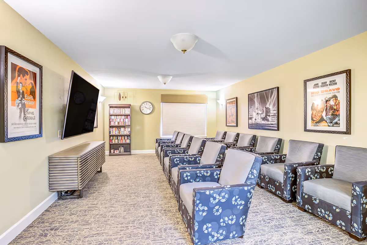 Bright communal lounge with a row of patterned armchairs facing a wall-mounted TV, bookshelves, and framed posters on the walls.