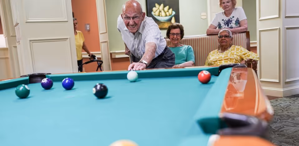 An elderly man is playing pool, aiming to hit the white cue ball on a green pool table. Three elderly women are watching him play, seated and standing nearby in a well-lit room with light-colored walls and a painting of pears on the wall.