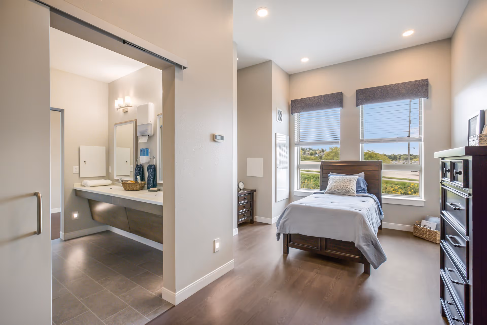 A bright and clean bedroom with a single bed made with gray bedding and a decorative pillow. The room has large windows with blinds partially open, allowing natural light to fill the space. There is a dark wooden dresser on the right side and a small nightstand with a clock near the bed. To the left, an open doorway leads to a bathroom area with a long countertop, two mirrors, and wall-mounted soap dispensers. The flooring is a combination of wood in the bedroom and tile in the bathroom.
